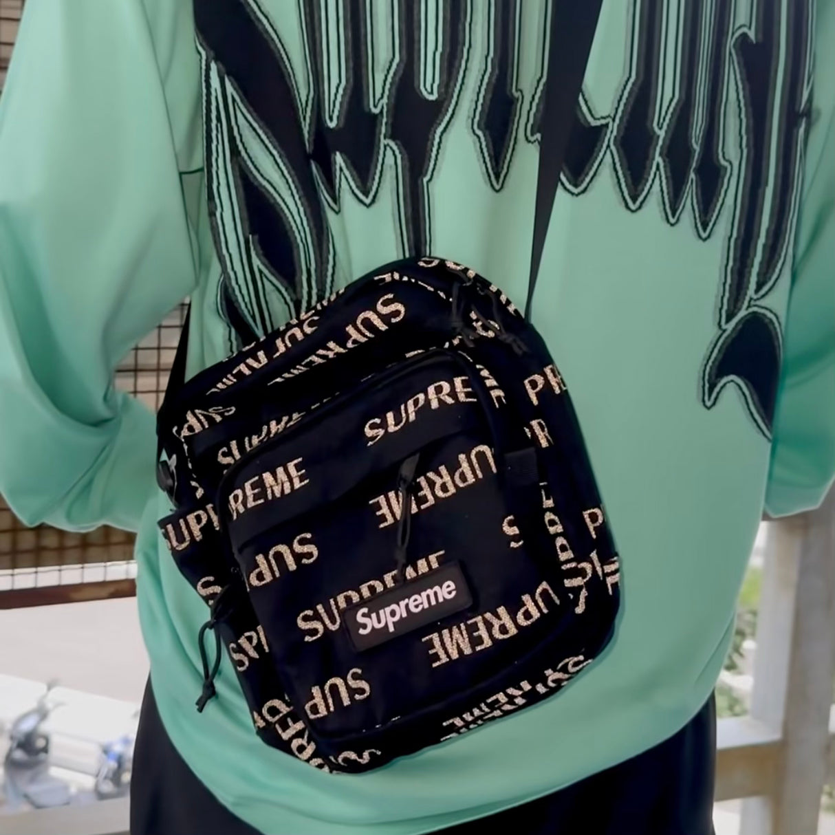Supreme black shoulder bag with reflective monogram logo, featuring a sleek design and adjustable strap.