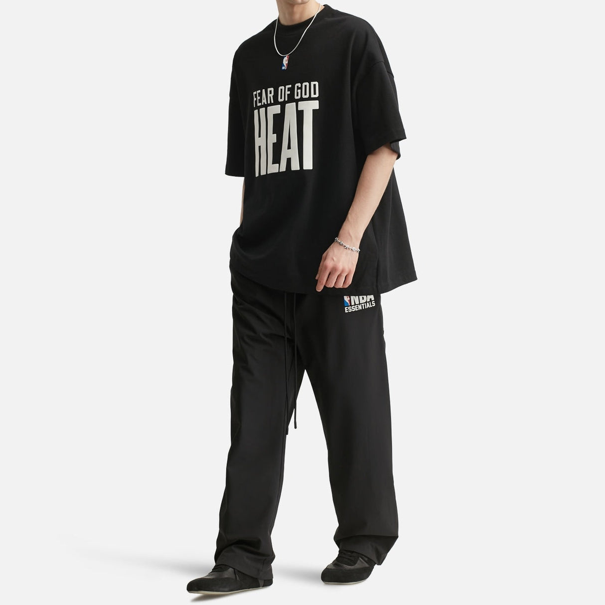 Fear of God Essentials NBA 90s Heavyweight Tee in black, featuring bold graphic design on a square image.